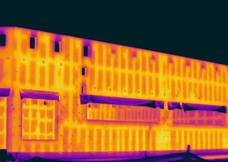 Night thermal image of a building facade. The wall glows orange with a regular vertical pattern of structural elements showing through the finish, while irregular cool purple areas appear around window heads and awnings where the pattern breaks.
