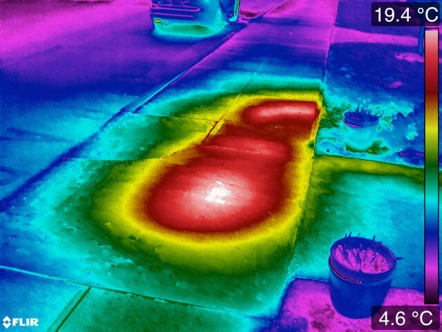 Thermal image of a tiled floor, scale 4.6 to 19.4 degrees Celsius. A large red and yellow plume of warmth spreads across several tiles from a point beneath the floor, with a sharp boundary into the cool blue-green of the surrounding dry slab.
