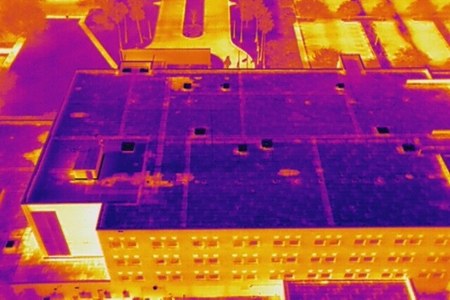 Aerial thermal image of a large low-slope commercial roof at dusk. The roof field reads cool where the assembly is dry, with warmer patches near curbs and drains, while surrounding pavement and the building facade read hot.